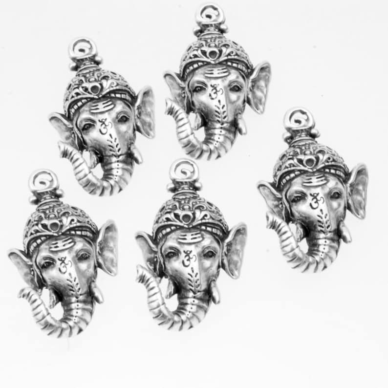 May include: Five silver-toned metal charms featuring the head of the Hindu deity Ganesha. Each charm has an intricate design and features the Om symbol on the forehead.