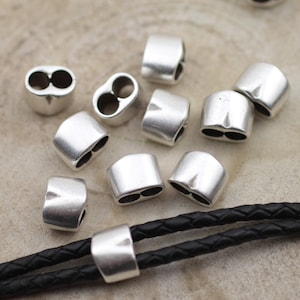 May include: Silver metal end caps for leather cord. The end caps have two holes for threading cord through. The end caps are shown on a black leather cord.