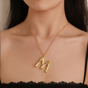 Puede incluir: Un collar de cadena dorada con un colgante de letra 'M' texturizado y grueso.