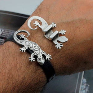 May include: A black leather bracelet with two silver gecko charms. One gecko charm has a detailed design and the other is more simple.