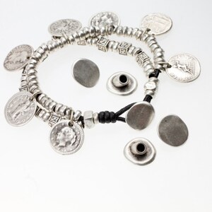 Pode incluir: Pulseira prateada com pingentes de moedas e contas decorativas. A pulseira é presa em um cordão preto e apresenta vários pingentes de moedas prateadas. Peças de metal prateadas adicionais também estão presentes.