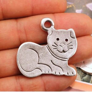 Op de afbeelding: Zilveren bedel in de vorm van een kat met een glimlachend gezicht. De bedel heeft een lus aan de bovenkant om aan een ketting of armband te bevestigen.