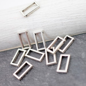 Puede incluir: Un grupo de anillos de salto rectangulares de metal plateado. Los anillos están dispersos sobre una superficie gris.