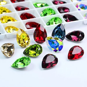 May include: Assortment of pear-shaped glass rhinestones in various colours, including red, green, blue, yellow, and gold. The rhinestones are arranged on a white surface.