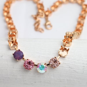 May include: A gold-coloured necklace featuring a series of round, colourful gemstones. The gemstones include shades of purple, pink, and iridescent colours. The necklace is displayed on a white surface.