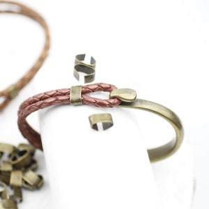 Peut inclure: Un bracelet en cuir marron avec une fermeture en m&eacute;tal et un design tress&eacute;. Le bracelet est sur une surface blanche avec d'autres fermoirs en m&eacute;tal.