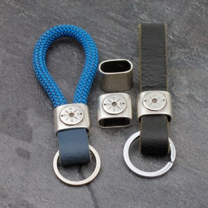 May include: Two keychains with compass charms. One keychain is made of blue rope and has a dark blue leather strap. The other keychain is made of black leather. Both keychains have silver-toned metal rings and compass charms.