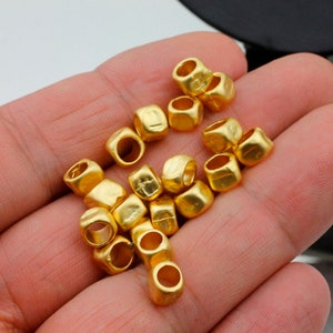 May include: A collection of gold-colored metal beads with a square shape and a hole in the center. The beads are scattered on a hand.