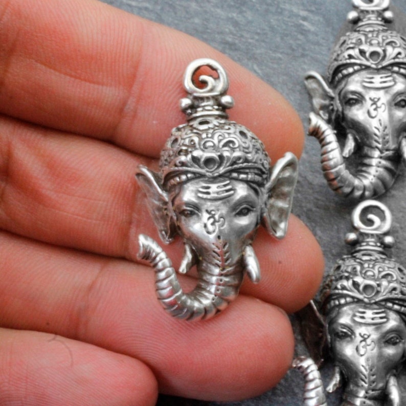 May include: Silver-toned metal pendant featuring a detailed depiction of the Hindu deity Ganesha, the elephant-headed god of wisdom and good fortune. The pendant has a traditional design with intricate carvings and a raised Om symbol on the forehead.