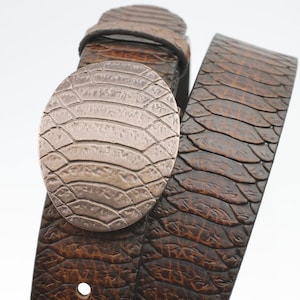 May include: A brown leather belt with a large, oval-shaped buckle. The buckle has a textured, embossed design that resembles a snakeskin pattern.