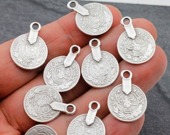 May include: Ten silver-toned metal coin charms with Arabic script and a star design. Each charm has a loop at the top for attaching to jewelry or other crafts.