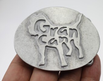 May include: Oval silver-coloured belt buckle featuring the words "Gran Can" and a dog silhouette. The buckle has a distressed finish, giving it a vintage appearance. The design is raised from the surface.