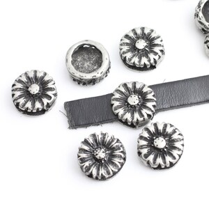 May include: Silver-toned metal flower-shaped charms with a small hole in the center. The charms are scattered on a white surface with a piece of black leather.