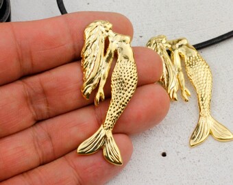 May include: Two gold-tone mermaid charms with detailed scales and flowing hair. The charms are hanging from a black leather cord.