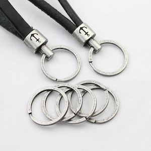 Può includere: Un portachiavi in ​​argento con un cinturino in pelle nera e un ciondolo a forma di ancora. Il portachiavi ha un anello grande e sei anelli più piccoli.