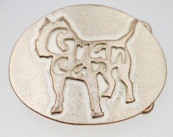 May include: A silver-coloured metal belt buckle in an oval shape. The design features a stylized dog silhouette with the words "Gran Can" cut out within the dog's form. The buckle has a brushed metal finish.