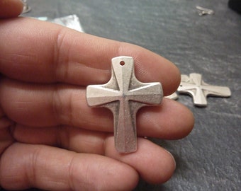 May include: A silver-colored cross pendant with a textured surface. The cross has a classic design with a hole at the top for a chain. Additional crosses are visible in the background. Religious jewelry.