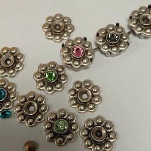 Puede incluir: Primer plano de varios adornos plateados en forma de flor con centros de piedras preciosas de colores. Las piedras preciosas son rosas, verdes y azules. Algunos de los adornos tienen un agujero en el centro.