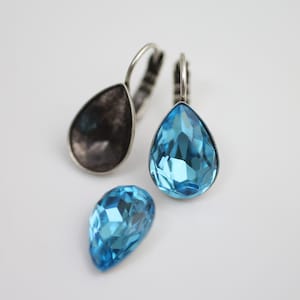 Puede incluir: Pendientes plateados con piedras preciosas azules en forma de lágrima. Un pendiente tiene una lágrima oscura y texturizada, mientras que el otro presenta una gema facetada de color azul claro. También hay una gema azul más pequeña suelta.
