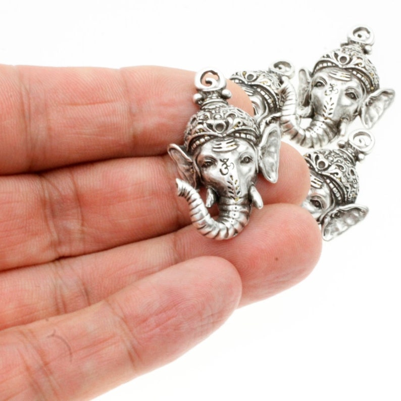May include: Four silver-toned metal charms featuring the head of the Hindu deity Ganesha. Each charm has intricate details and a raised Om symbol on the forehead.