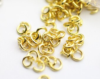 May include: Close-up of gold-colored jewelry findings, including open jump rings and connectors. The findings are scattered on a white surface. These small, shiny components are used in jewelry making and crafting.