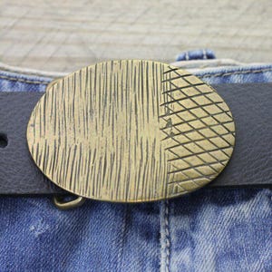 Peut inclure: Boucle de ceinture ovale en laiton avec un motif texturé. La boucle est fixée à une ceinture en cuir noir, sur fond de jean bleu. La boucle présente un motif vertical et en losange.