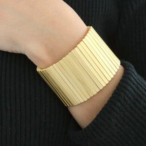 May include: A wide, gold-colored cuff bracelet. The bracelet is made of numerous vertical rectangular bars, creating a textured, geometric design. The jewelry is worn on a wrist, against a dark background.