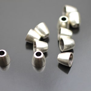 May include: A group of silver metal cord end caps, also known as cord tips, are scattered on a gray surface. The cord ends are round and have a slight dome shape.