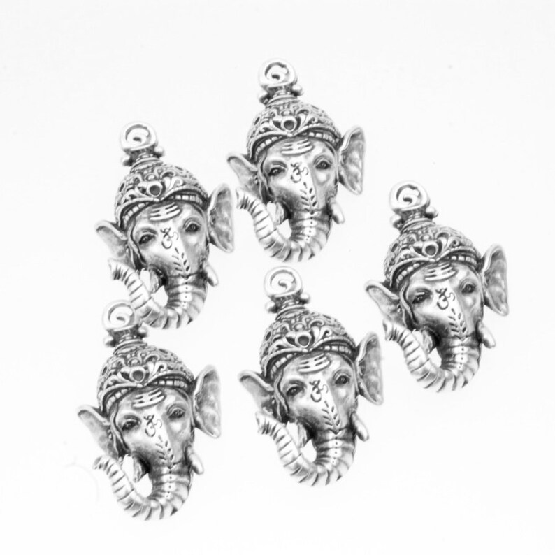 May include: Five silver-toned metal charms featuring the face of the Hindu deity Ganesha, the elephant-headed god of wisdom and good fortune. Each charm has intricate details and a loop for attaching to jewelry or other crafts.