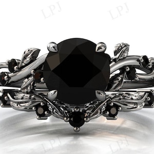 Puede incluir: Un anillo de compromiso de diamante negro con un delicado diseño floral. El anillo presenta un gran diamante negro redondo rodeado de diamantes negros más pequeños y detalles intrincados de hojas. La banda es de plata y tiene un acabado pulido.