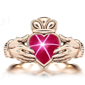 Op de afbeelding: Een ring van goudkleurig metaal met een rode, hartvormige steen in een Keltisch knoopontwerp. De ring toont twee handen die het hart vasthouden, wat liefde en eenheid symboliseert.