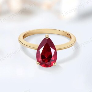 Op de afbeelding: Een gouden ring met een peervormige, dieprode edelsteen. De ring heeft een gepolijste gouden band en de edelsteen wordt op zijn plaats gehouden door drie kleine pootjes. Het ontwerp is elegant en klassiek, geschikt voor diverse gelegenheden.