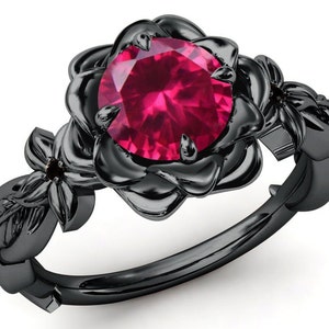 May include: Black rose engagement ring featuring a large, round, red gemstone in the centre. The ring is crafted from black metal and showcases intricate floral details.