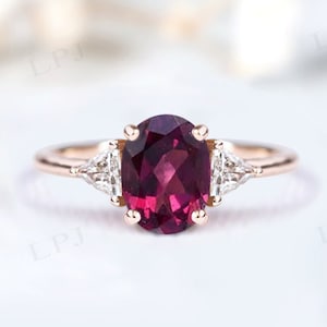 May include: A rose gold engagement ring featuring a large oval ruby gemstone, flanked by two smaller triangular diamonds.