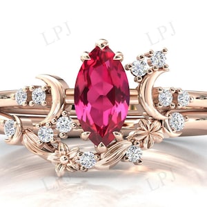 May include: A rose gold ring set with a large, marquise-shaped ruby gemstone. The ring features a delicate floral design with small, clear gemstones and crescent moon accents.