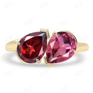 May include: A gold ring with two pear-shaped gemstones. The left gemstone is a deep red garnet, and the right gemstone is a pink topaz.