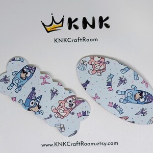 May include: Two blue and pink patterned hair clips featuring a cartoon dog wearing a party hat. The clips are on a white background with the text "KNK Craft Room" and "www.KNK Craft Room.etsy.com".