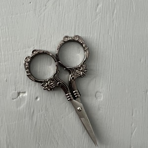 May include: Silver-toned decorative scissors with ornate handles and a pointed blade. The scissors are on a white textured surface.