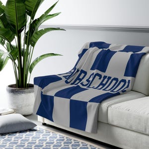 May include: A blue and white checkered throw blanket draped over a white couch. The blanket features the text "OUR SCHOOL" in large, bold letters. A large potted plant is in the background.