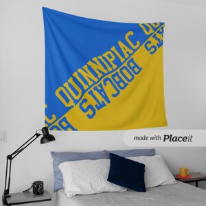 May include: A blue and gold tapestry with the text "Quinnipiac Bobcats" in yellow lettering. The tapestry is hanging on a white wall above a bed with white bedding and pillows. A black lamp and side table are visible.