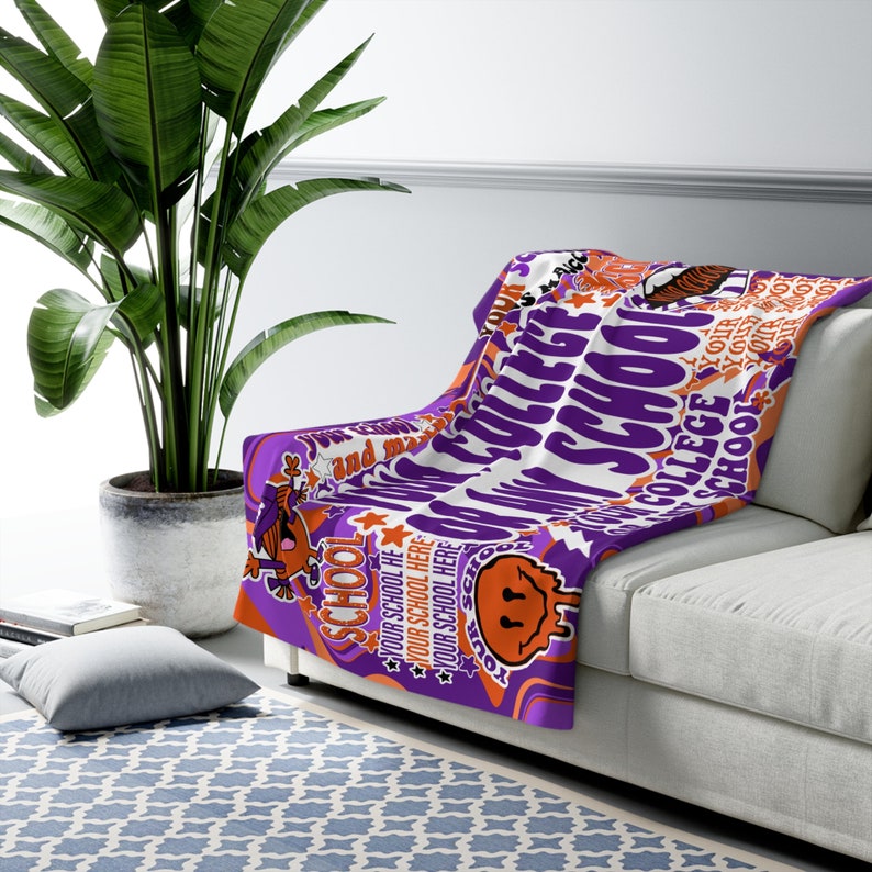 Peut inclure: Une couverture de jet&eacute; d'&eacute;cole universitaire violette et blanche drap&eacute;e sur un canap&eacute; blanc. La couverture pr&eacute;sente les mots "COLLEGE SCHOOL" et "YOUR SCHOOL HERE" dans un design r&eacute;tro avec des accents orange et un smiley.