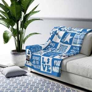 May include: A white and blue throw blanket with a repeating pattern of squares and text. The text includes phrases like "Your School" and "Saturdays are for Mascot". The blanket is draped over a white couch.