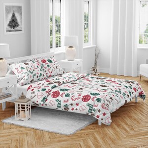 Op de afbeelding: Wit kerstthema beddengoed met rode, groene en bruine feestelijke patronen. De set bevat een dekbedovertrek en twee kussenslopen, met zuurstokken, kerstballen en sterren. Het bed staat in een lichte slaapkamer met witte meubels.