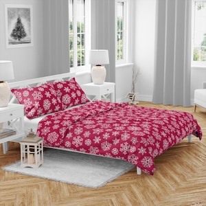 Op de afbeelding: Een rode dekbedovertrekset met een wit sneeuwvlokpatroon. De set bevat een dekbedovertrek en twee kussenslopen. Het beddengoed ligt op een wit bedframe in een slaapkamer. De kamer heeft witte gordijnen en een houten vloer.