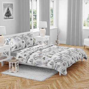 Op de afbeelding: Een wit bed met een winterthema dekbedovertrek en kussenset. Het beddengoed heeft een patroon van grijze en witte bomen en andere winterelementen. De kamer heeft witte meubels, grijze gordijnen en een ingelijste foto van een boom.