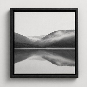 May include: Black and white framed canvas art print featuring a serene landscape. The artwork depicts a mountain range reflected in a calm lake, with fog partially obscuring the peaks. The frame is a dark, solid color.