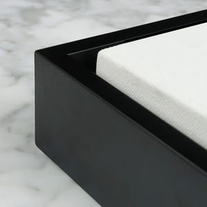 May include: A black rectangular box with a white, textured interior. The box has a matte finish and a simple, modern design. The corner of the box is visible, showcasing the clean lines and sharp angles. The box appears to be empty.