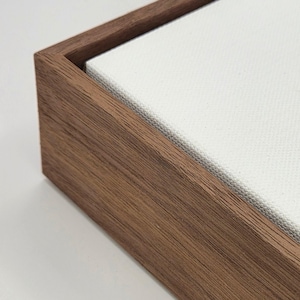 May include: Close-up of a wooden frame with a white canvas. The frame is made of dark brown wood with visible grain. The canvas is stretched tightly within the frame, creating a clean, modern look. The corner of the frame is visible.