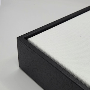 May include: Close-up of a black wooden frame with a white canvas stretched inside. The frame has a dark, textured finish. The canvas is tightly stretched, creating a clean, modern look. Ideal for artwork or photography.