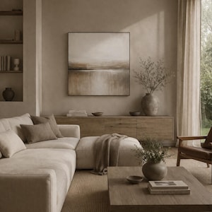 May include: A neutral-toned living room with a large abstract painting above a wooden cabinet. A beige sectional sofa with throw pillows and a wooden coffee table are in the foreground. A large vase with branches sits on the cabinet.
